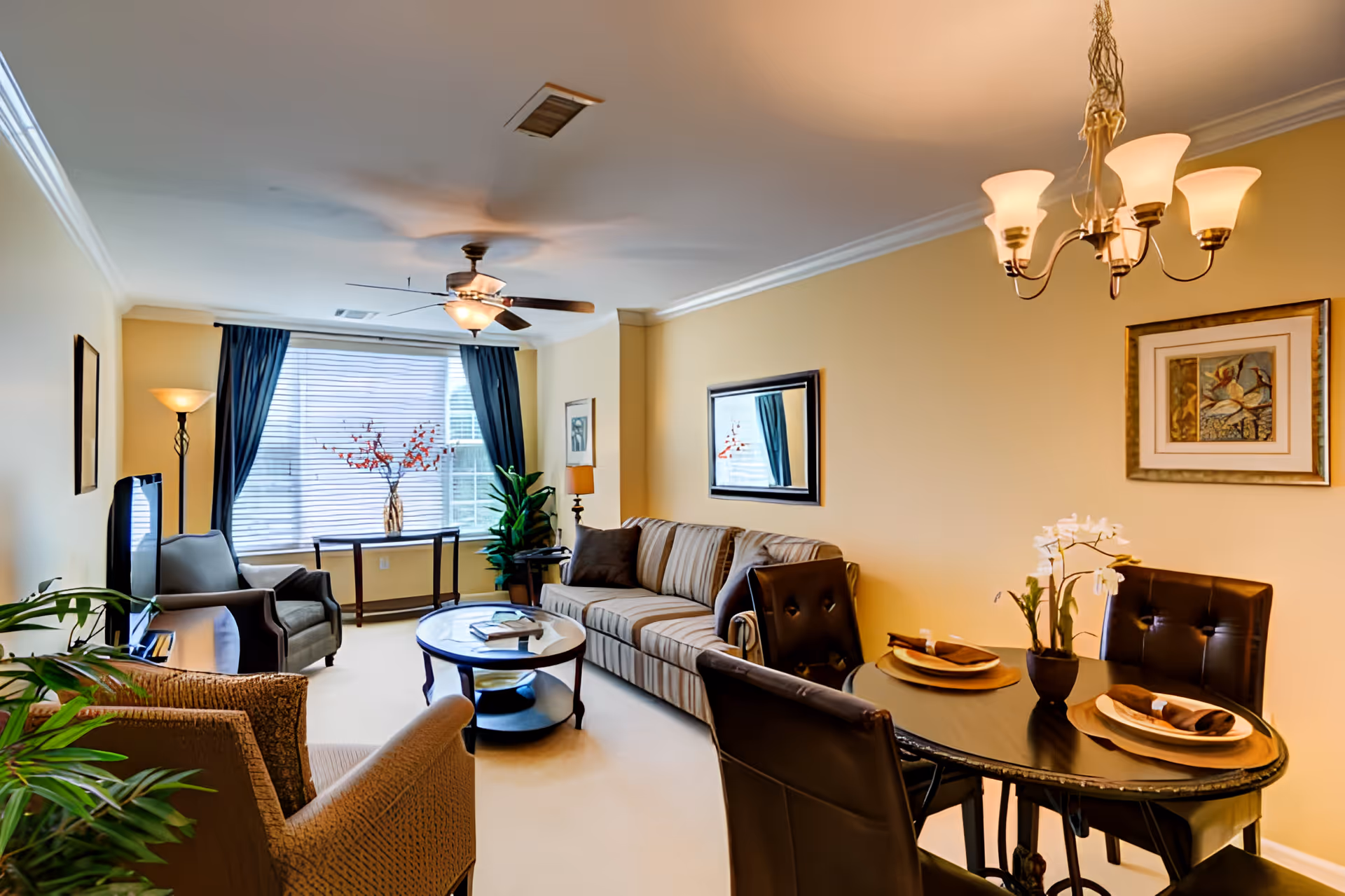 Bright furnished living room with a sofa, armchairs, coffee table and a small dining table beside a large window with blue curtains.