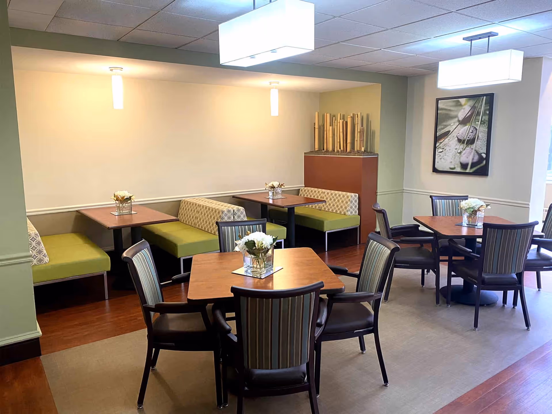 Well-lit dining area with wooden tables, striped chairs, green cushioned booths, and small floral centerpieces.