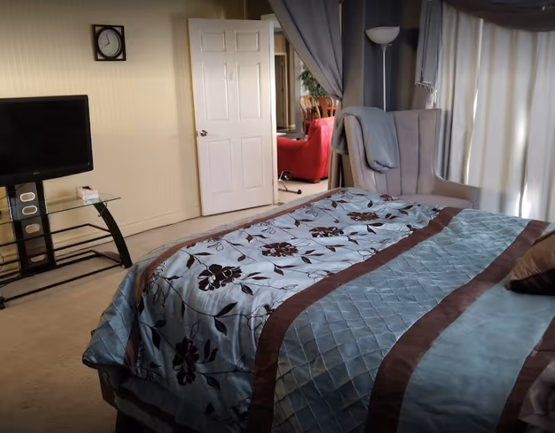 A bedroom with a bed covered in a blue and brown floral patterned comforter. There is a gray armchair with a blanket draped over it near a window with curtains. A flat-screen TV is mounted on a glass stand against the wall, and a clock is hanging above it. The door is open, showing a glimpse of a red chair and a dining area beyond.