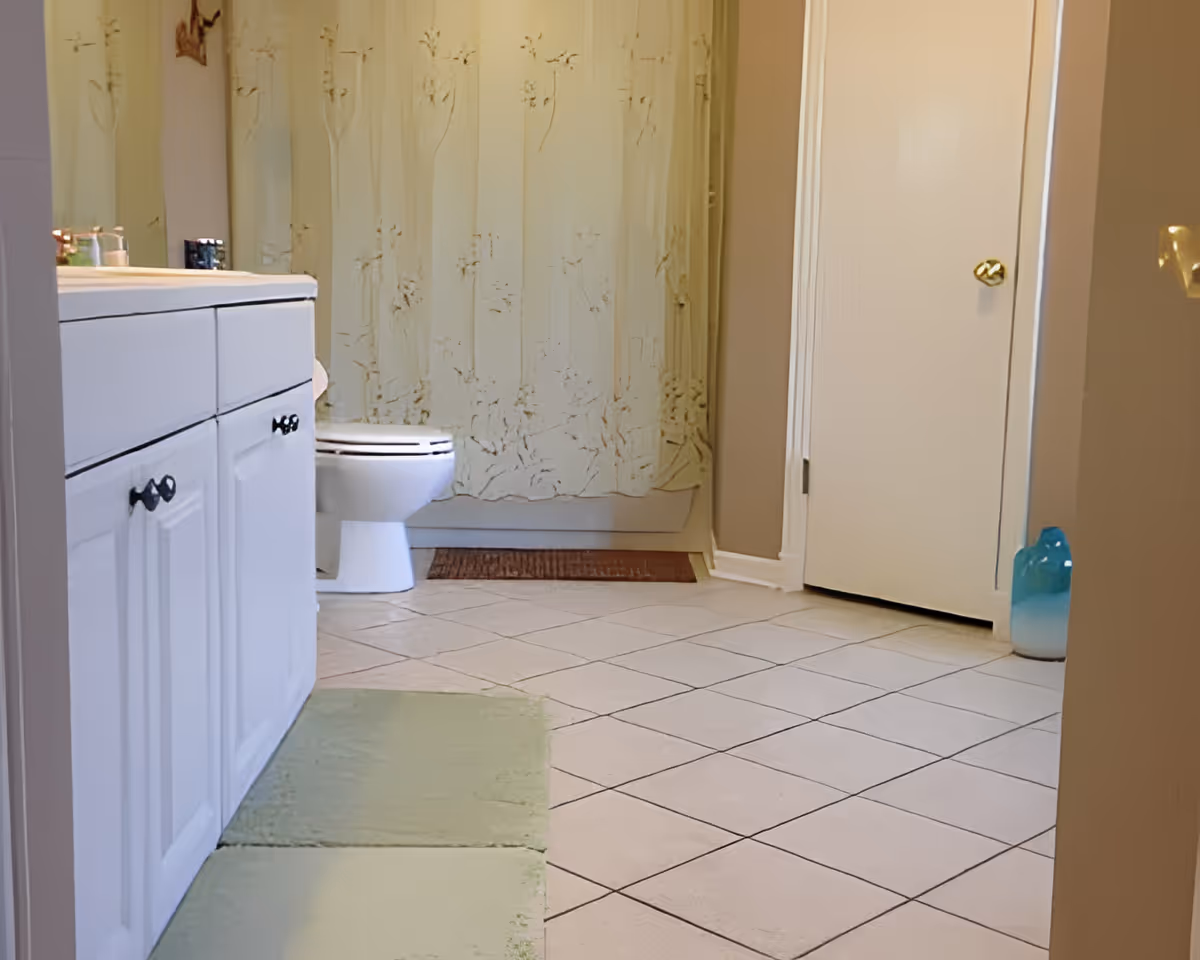 Bright bathroom with a white vanity and toilet, patterned shower curtain, tiled floor, and green bath mats.