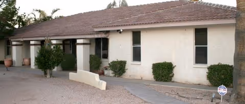 Single-story building with a tiled roof and beige exterior walls, featuring several windows and a walkway leading to the entrance, surrounded by small bushes and trees.