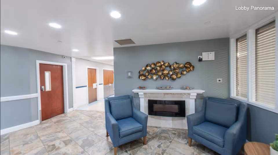 A lobby area with two blue armchairs facing a decorative fireplace with a metallic wall art above it. The floor is tiled, and there are several wooden doors along a hallway. Windows with blinds are on the right side, and the walls are painted in a soft gray color. The image has the text 'Lobby Panorama' in the top right corner.