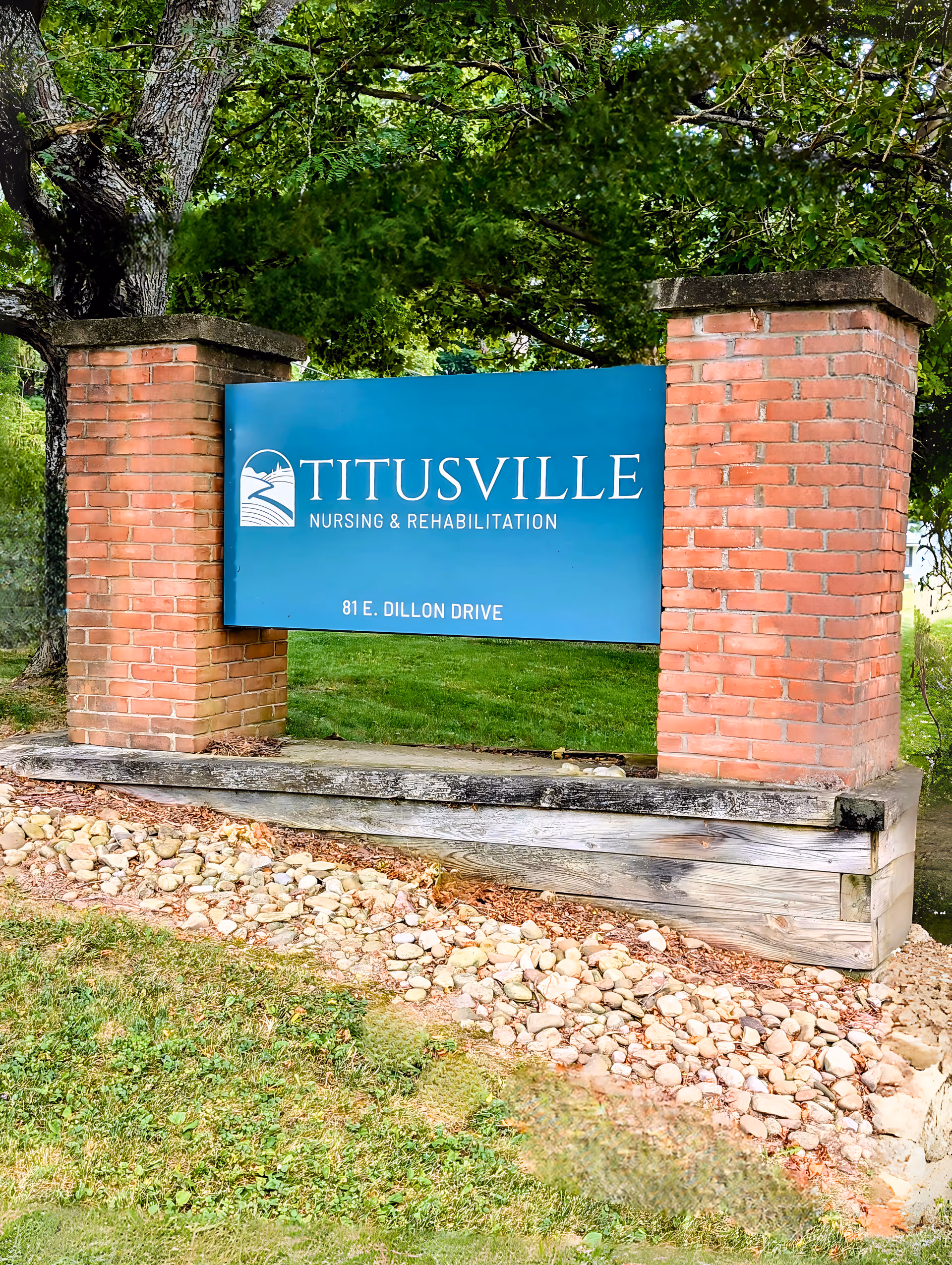 A blue sign reading "TITUSVILLE Nursing & Rehabilitation, 81 E. Dillon Drive" mounted between two brick pillars with grass and trees behind.