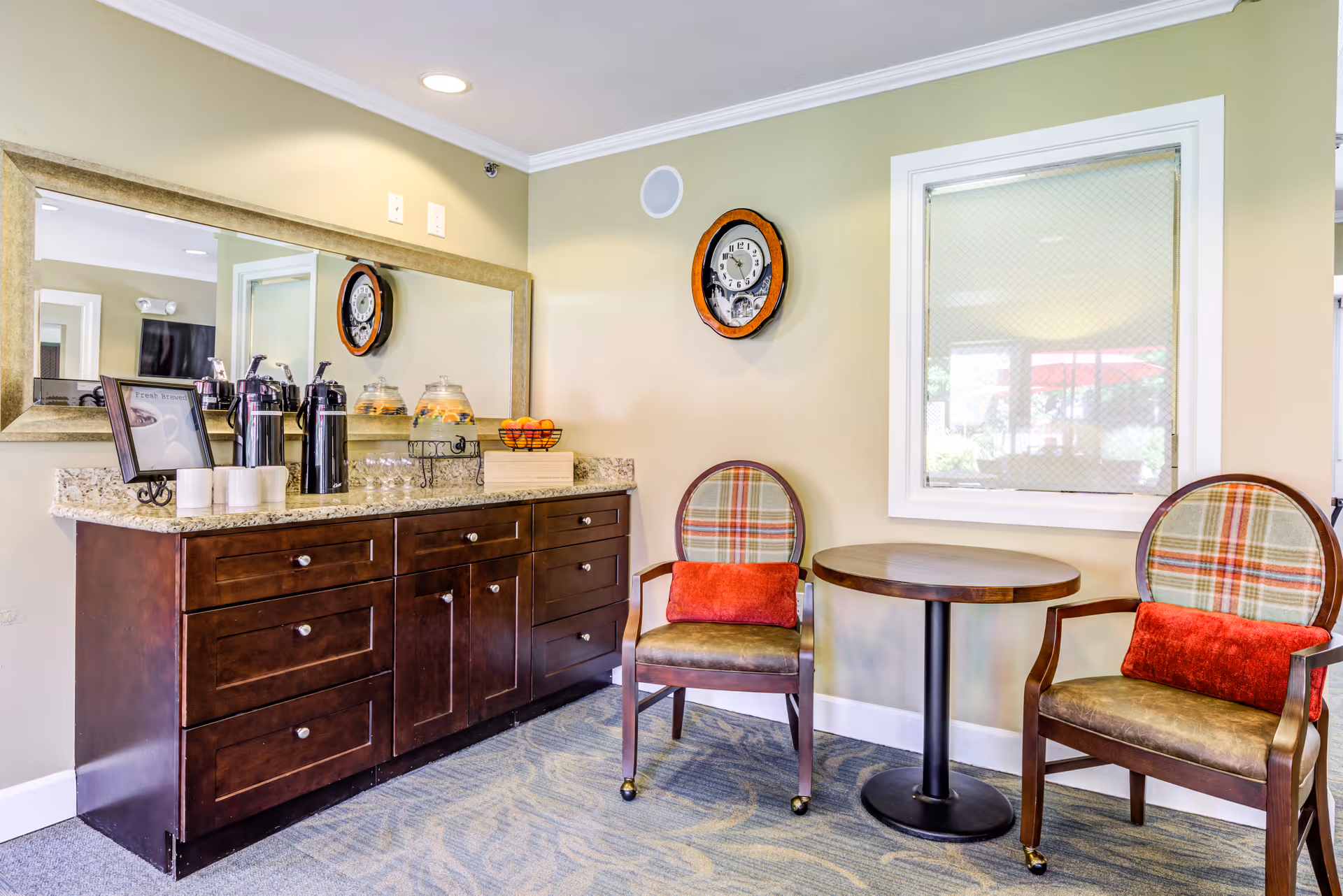 Two upholstered chairs with red cushions and a small round table next to a countertop with coffee dispensers and snacks in a bright common area.