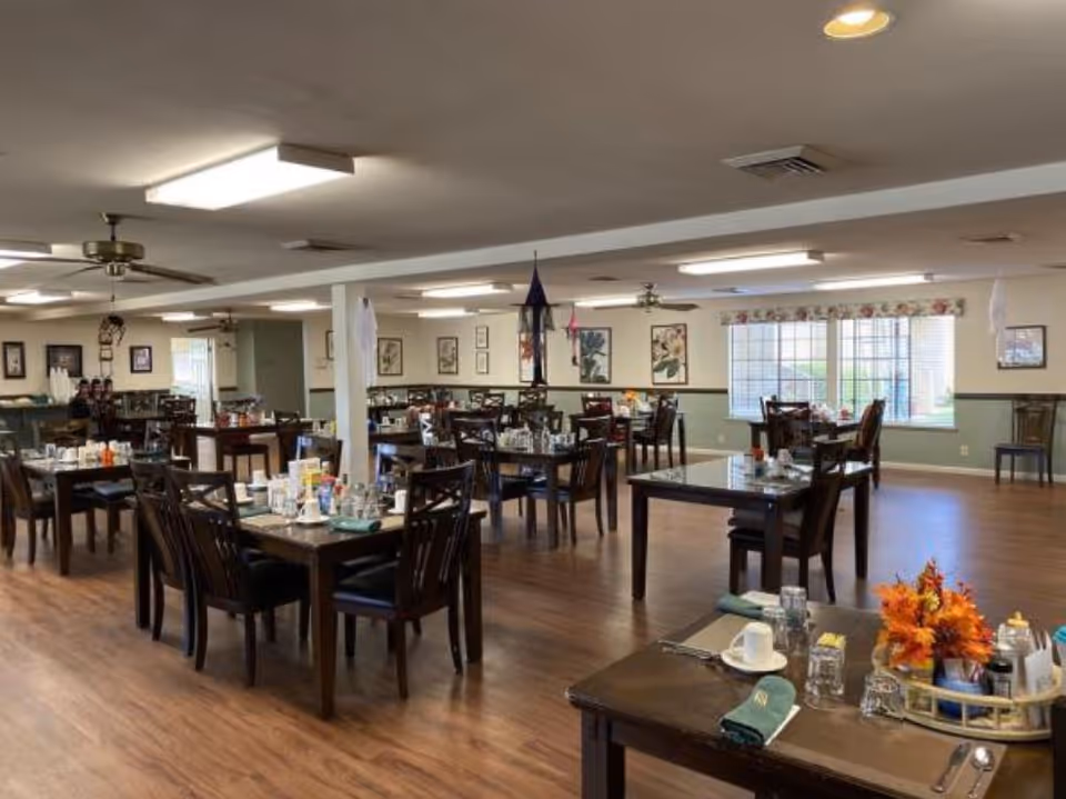 A spacious dining room in a retirement community with multiple dark wooden tables and chairs arranged neatly. Each table is set with glasses, napkins, and condiments. The room has wooden flooring, ceiling fans, fluorescent lighting, and large windows with floral valances allowing natural light to enter. The walls are decorated with framed pictures.