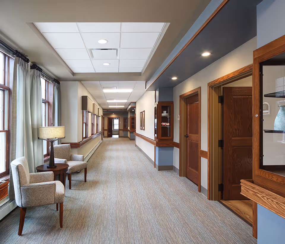 A well-lit hallway in an assisted living facility with large windows on the left side covered by light curtains. Two upholstered chairs and a small round table with a lamp are placed near the windows. The hallway has carpeted flooring, wooden trim, and several wooden doors on the right side. Display cabinets with decorative items are mounted on the walls.