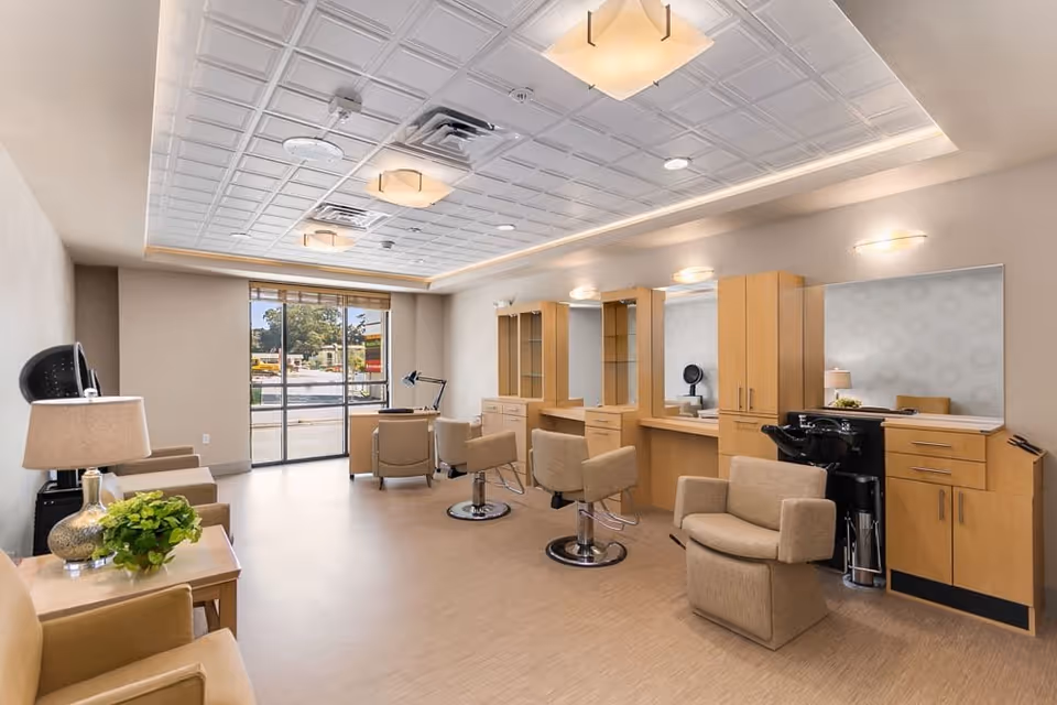 A bright and spacious salon area with beige chairs, wooden cabinetry, large mirrors, and a hair washing station. The room has a modern ceiling with recessed lighting and large windows letting in natural light.