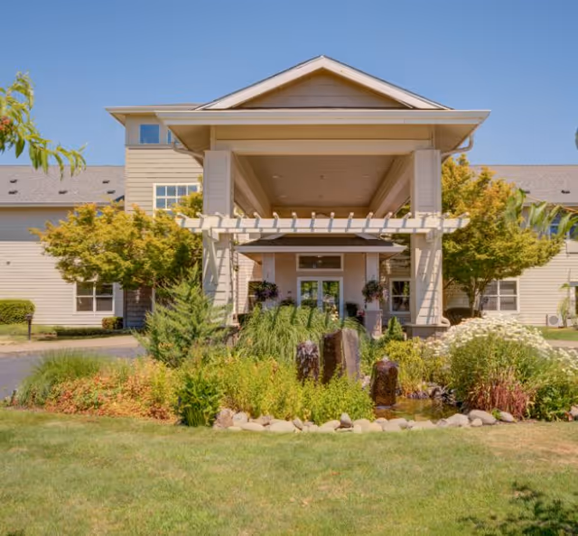 Front exterior view of a senior living facility with a covered entrance, surrounded by well-maintained landscaping including green shrubs, trees, and a small water feature with rocks. The building is light-colored with multiple windows and a clear blue sky above.
