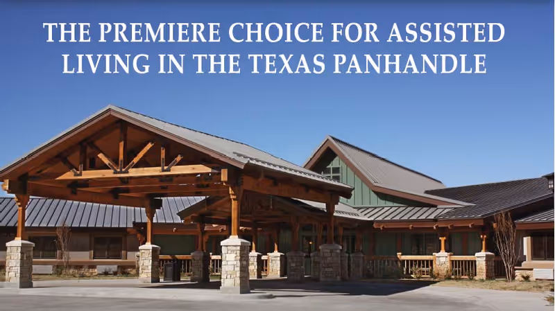 Front entrance of an assisted living facility with a timber-framed covered porte-cochere and stone pillars under a clear blue sky.