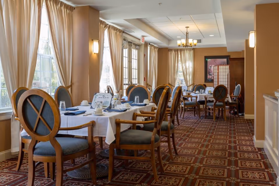 A dining room with several tables covered with white tablecloths, set with cups, glasses, and napkins. The room has large windows with beige curtains, wooden chairs with blue cushions, and a patterned carpet. Warm lighting fixtures are mounted on the walls and ceiling.