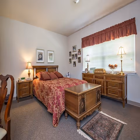 A cozy bedroom with a wooden bed frame and a red patterned bedspread, matching pillows, a wooden nightstand with a lamp, a wooden chest at the foot of the bed, a wooden dresser with two lamps and decorative items, a window with blinds and a red valance, framed pictures on the walls, and a small rug on the carpeted floor.