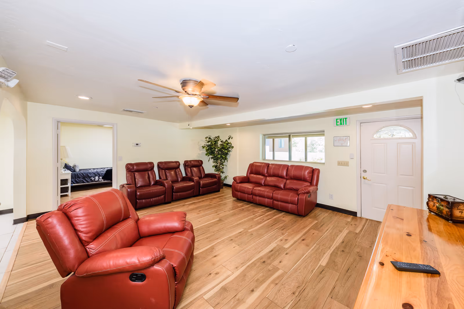 A bright living room with wooden flooring, featuring a red leather armchair in the foreground, a matching red leather sofa and three recliners against the far wall. There is a ceiling fan with a light fixture, a potted plant in the corner, a window, and a white door with an exit sign above it. A wooden table with a remote control and a decorative box is visible on the right side. An adjacent room with a bed and nightstand is visible through an open doorway.