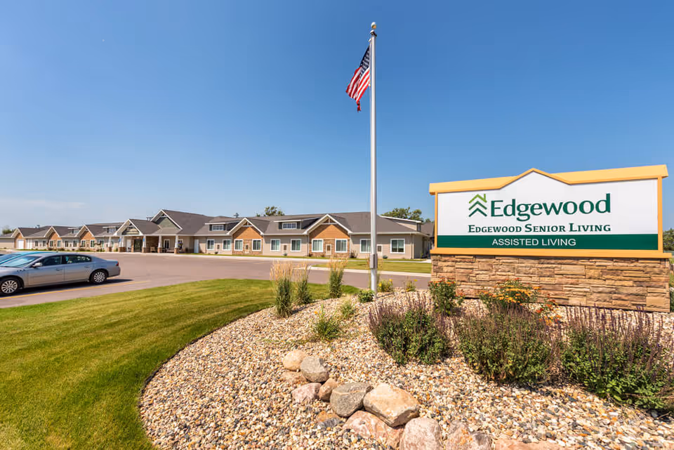 Exterior view of Edgewood Mitchell senior living facility on a clear sunny day, showing a single-story building with multiple entrances, a parking area with cars, a landscaped area with rocks and plants, and an American flag on a flagpole near a large sign that reads 'Edgewood Senior Living Assisted Living'.