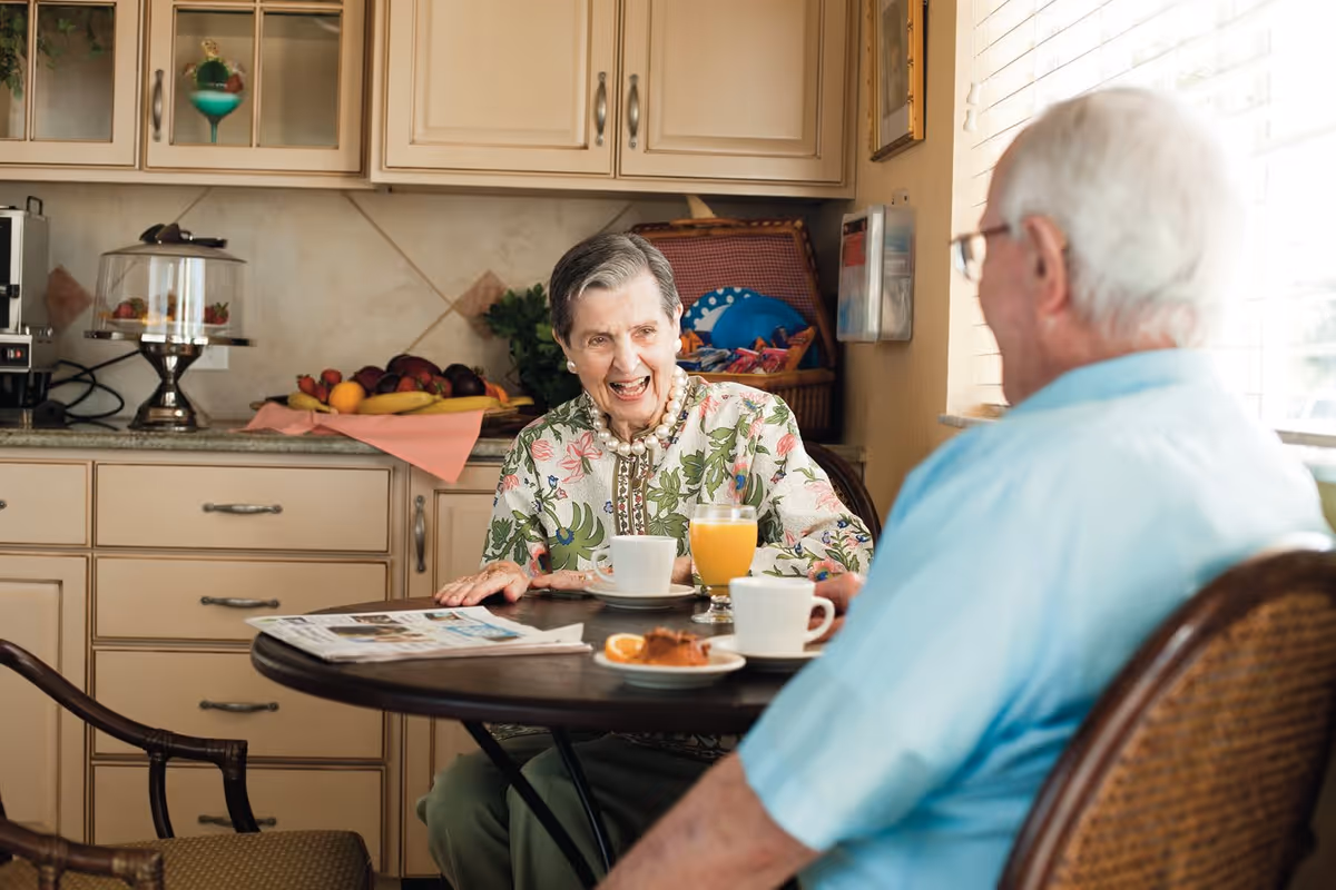 An elderly woman and man sitting at a round table in a kitchen or dining area, enjoying a conversation. The woman is smiling and wearing a floral blouse, while the man is seen from behind wearing a light blue shirt. On the table are cups of coffee, a glass of orange juice, a pastry, and a newspaper. The background shows kitchen cabinets, a fruit basket, and a coffee machine.