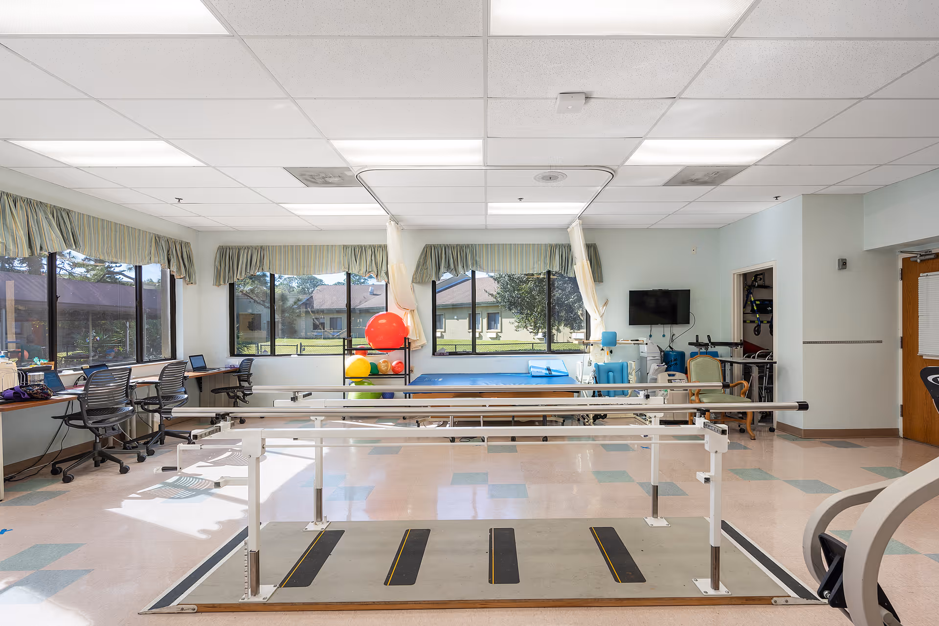 A bright rehabilitation room with parallel bars for walking exercises in the center. The room has large windows with green valances, letting in natural light and showing a view of a grassy outdoor area. There are exercise balls and therapy equipment near the windows, a blue therapy table, a wall-mounted TV, and several chairs and desks along the walls.