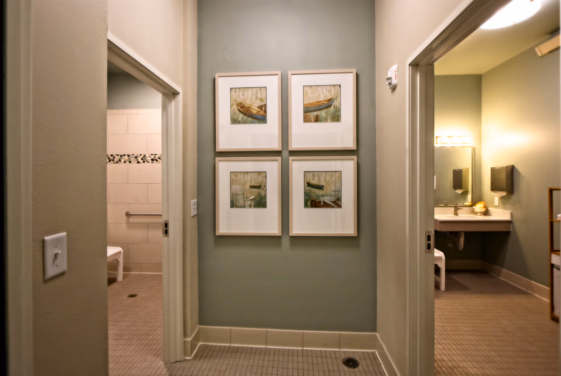 A tiled hallway with four framed prints on the center wall and open doorways to accessible bathrooms on either side.