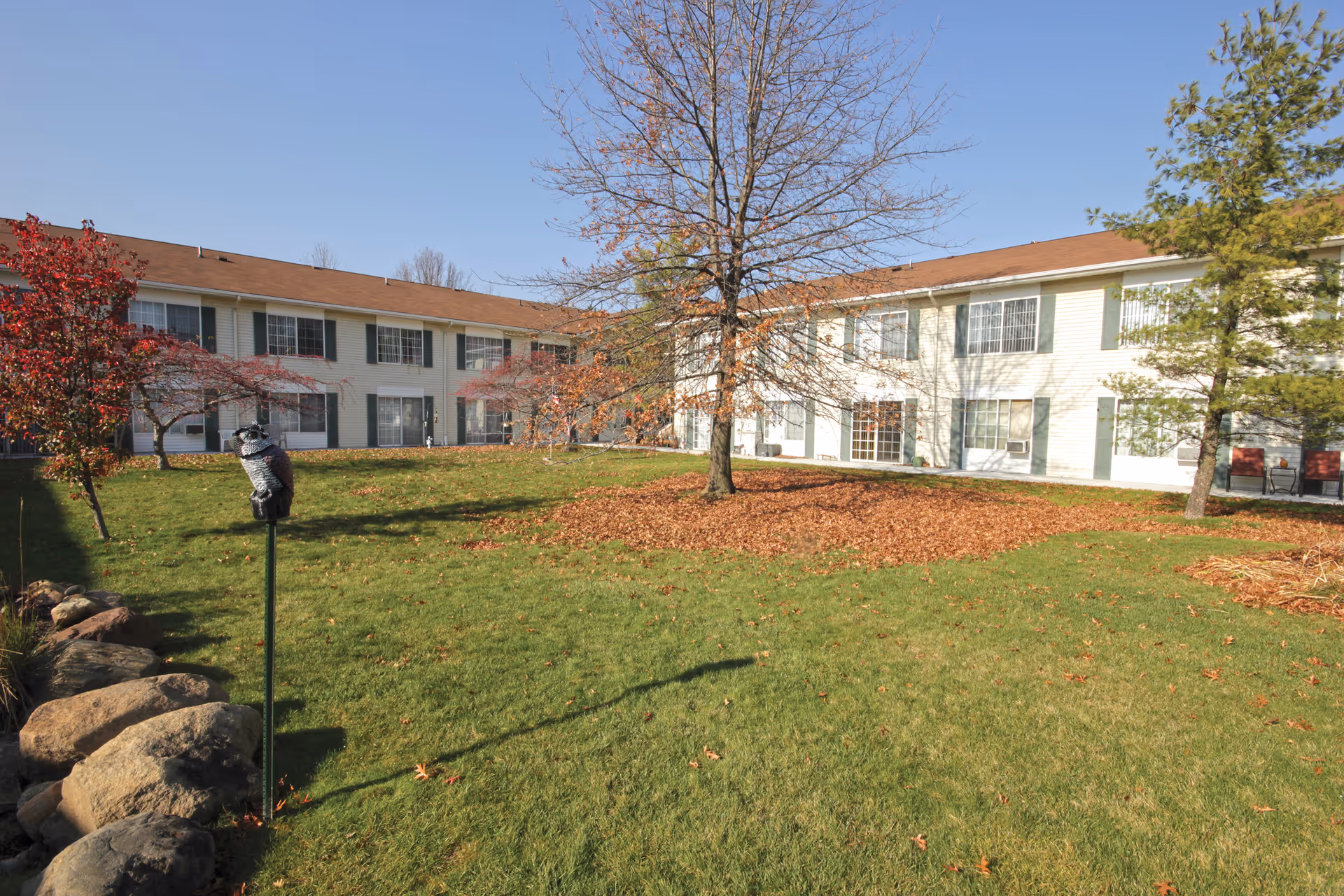 Two-story senior living building with a grassy courtyard, trees with fallen leaves, and a stone border under a clear sky.