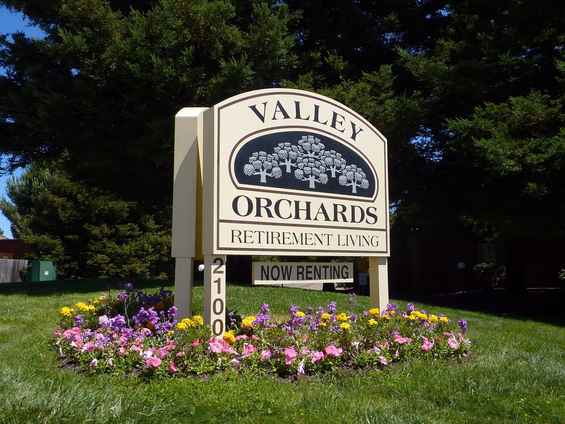 A large sign for Valley Orchards Retirement Living stands on a grassy area surrounded by colorful flowers, with tall green trees in the background. The sign includes the text 'VALLEY ORCHARDS RETIREMENT LIVING' and a smaller sign below that reads 'NOW RENTING'.