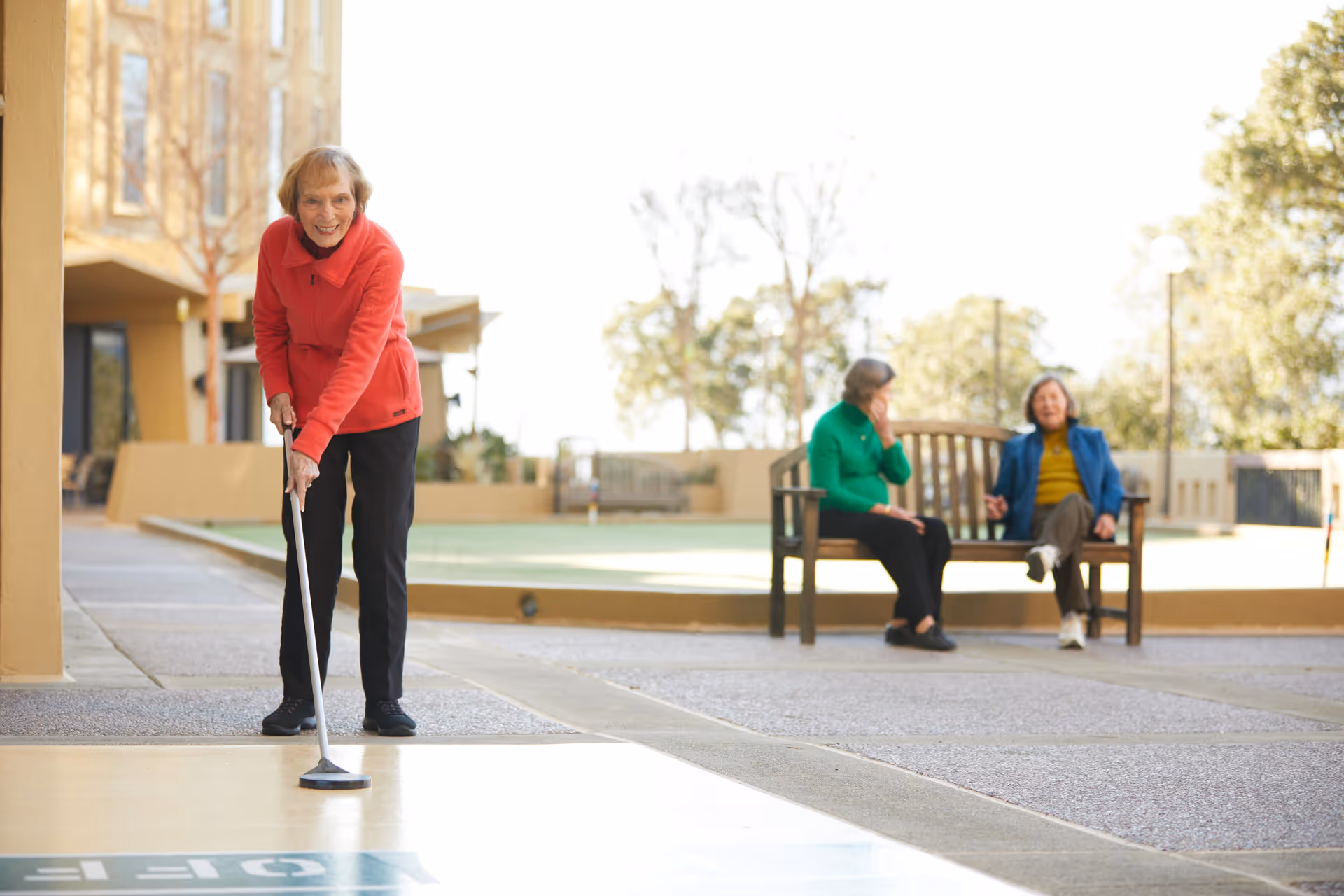 An elderly woman in a red jacket playing shuffleboard outdoors on a sunny day, with two other elderly women sitting and chatting on a bench in the background near a building.