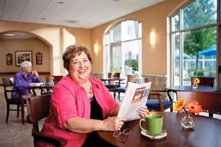 A smiling elderly woman in a bright pink jacket sitting at a table in a dining area, holding a newspaper. There is a green coffee cup and a small vase with orange flowers on the table. In the background, another elderly woman is seated at a different table near large windows letting in natural light.