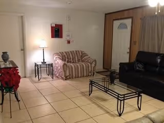 A living room with tiled floor featuring a striped loveseat, a black leather sofa, a glass coffee table with a decorative item, a side table with a lamp, and a small table with a vase of red flowers. There is a white door and a window with beige curtains in the background.