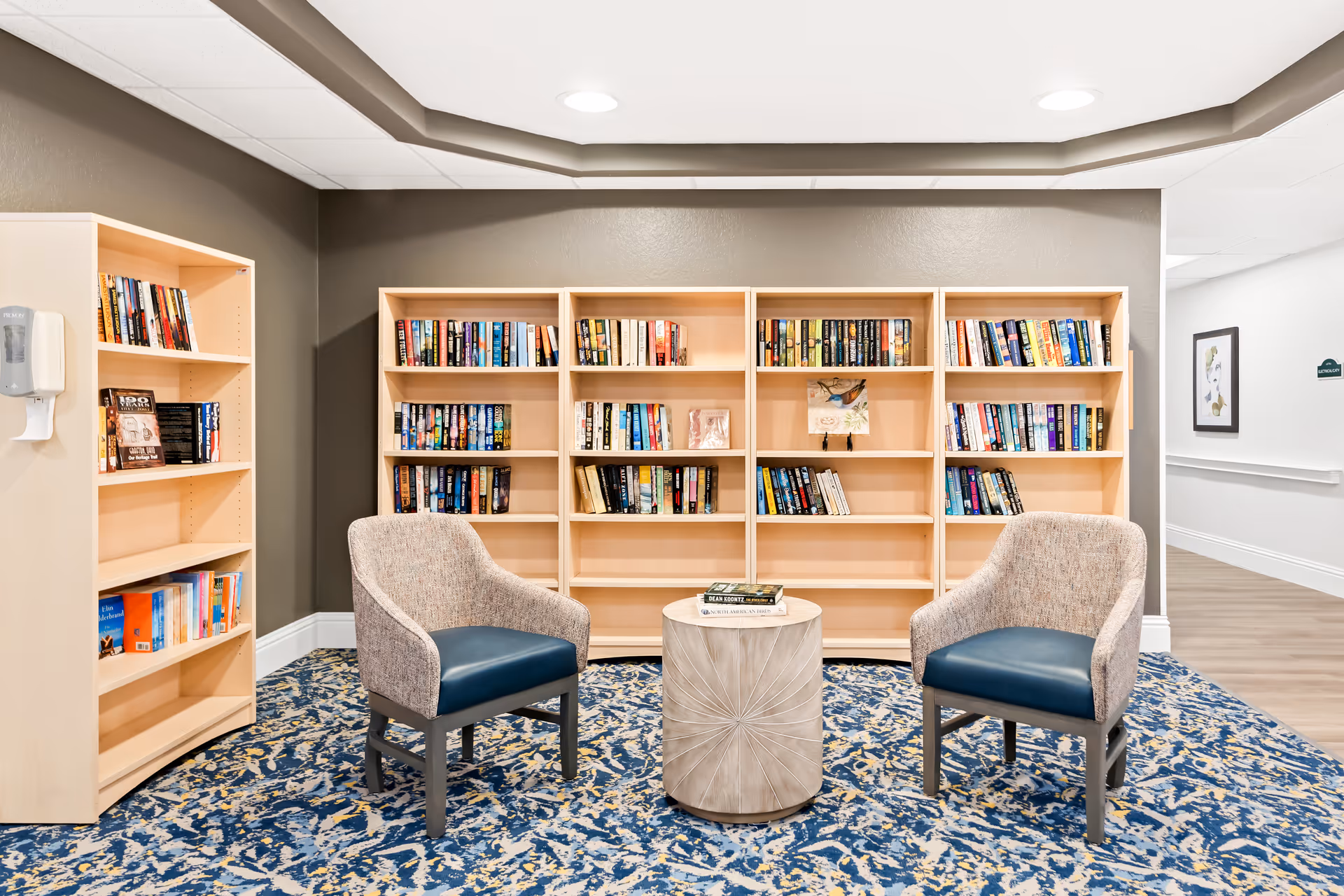 A cozy reading nook in a senior living facility featuring two upholstered armchairs with blue seats positioned around a small round wooden table. Behind the chairs are light wood bookshelves filled with various books. The area has a patterned blue and beige carpet, gray walls, and a white ceiling with recessed lighting. A hallway with white walls and framed artwork is visible to the right.