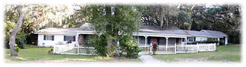 Exterior view of a single-story residential building with a white picket fence, surrounded by trees and greenery under a bright sky.