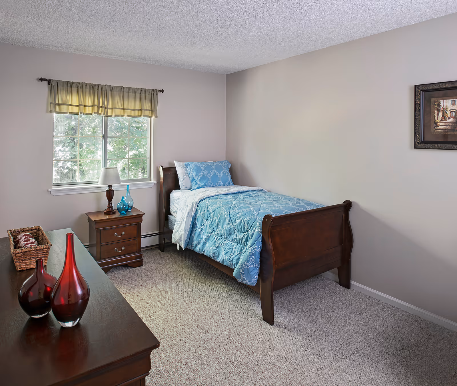 A simple bedroom with a single wooden bed covered with a blue patterned comforter and pillow. Next to the bed is a wooden nightstand with a lamp and two blue glass vases. A window with a light green valance lets in natural light. Across from the bed is a wooden dresser with two red glass vases and a basket. A framed picture hangs on the beige wall.