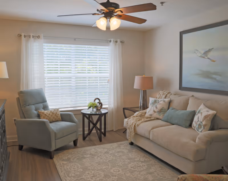 A cozy living room with a beige sofa adorned with floral and solid pillows, a light blue armchair with a patterned pillow, a round wooden side table with a small plant and decorative item, a floor lamp, a ceiling fan with lights, and a large window with white curtains letting in natural light. A framed painting of a bird in flight hangs on the wall above the sofa.