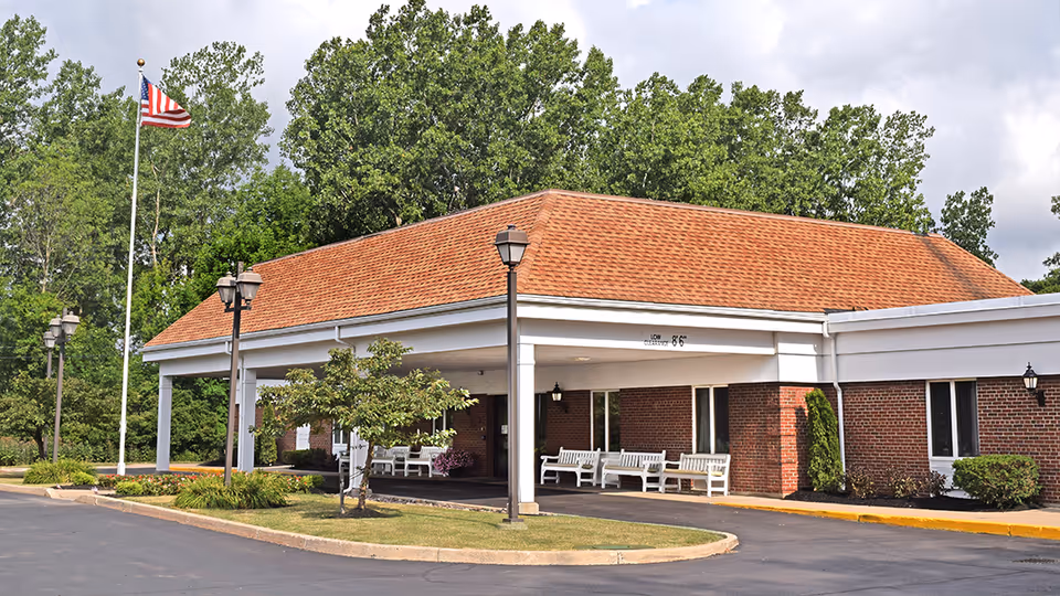 Covered entrance and porte-cochere of a single-story brick senior living building with benches, lamp posts, and an American flag.