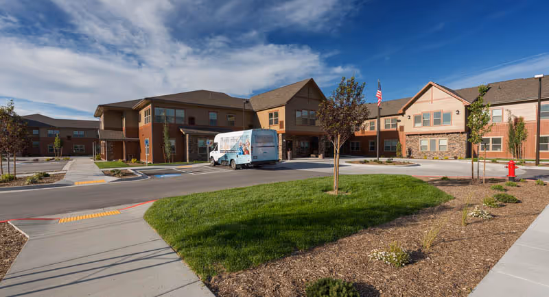 Exterior view of Summit Senior Living facility showing a large two-story building with a mix of brick and siding. There is a driveway with a parked shuttle bus, a flagpole with an American flag, landscaped grass and small trees, and a clear blue sky with some clouds.
