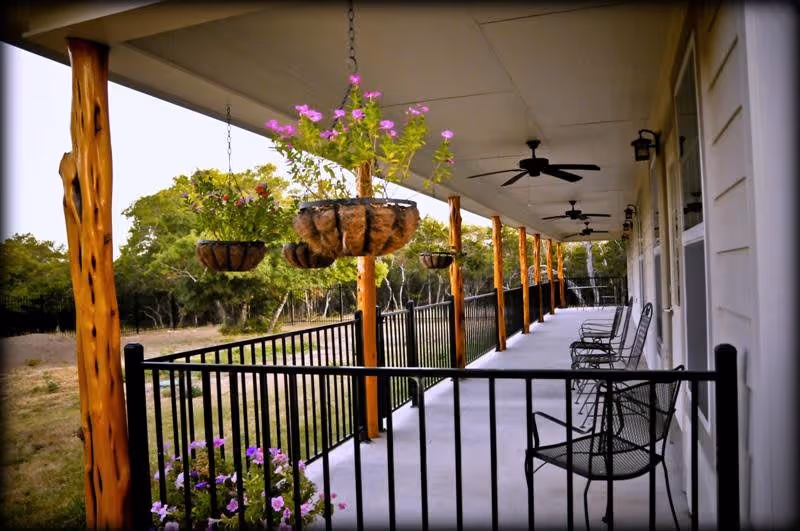 A covered outdoor porch area with wooden posts and black metal railing. Hanging baskets with pink flowers are suspended from the ceiling. Several black metal chairs are arranged along the porch. Ceiling fans are mounted overhead. Trees and greenery are visible in the background.