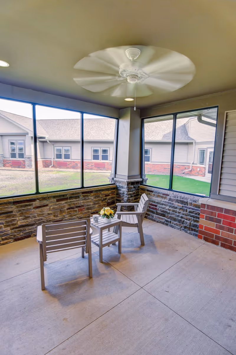 A screened-in porch area with two gray chairs and a small table between them holding a vase of yellow flowers. The porch has stone and brick half walls and large windows looking out to a grassy courtyard and building exterior. A ceiling fan is spinning above.