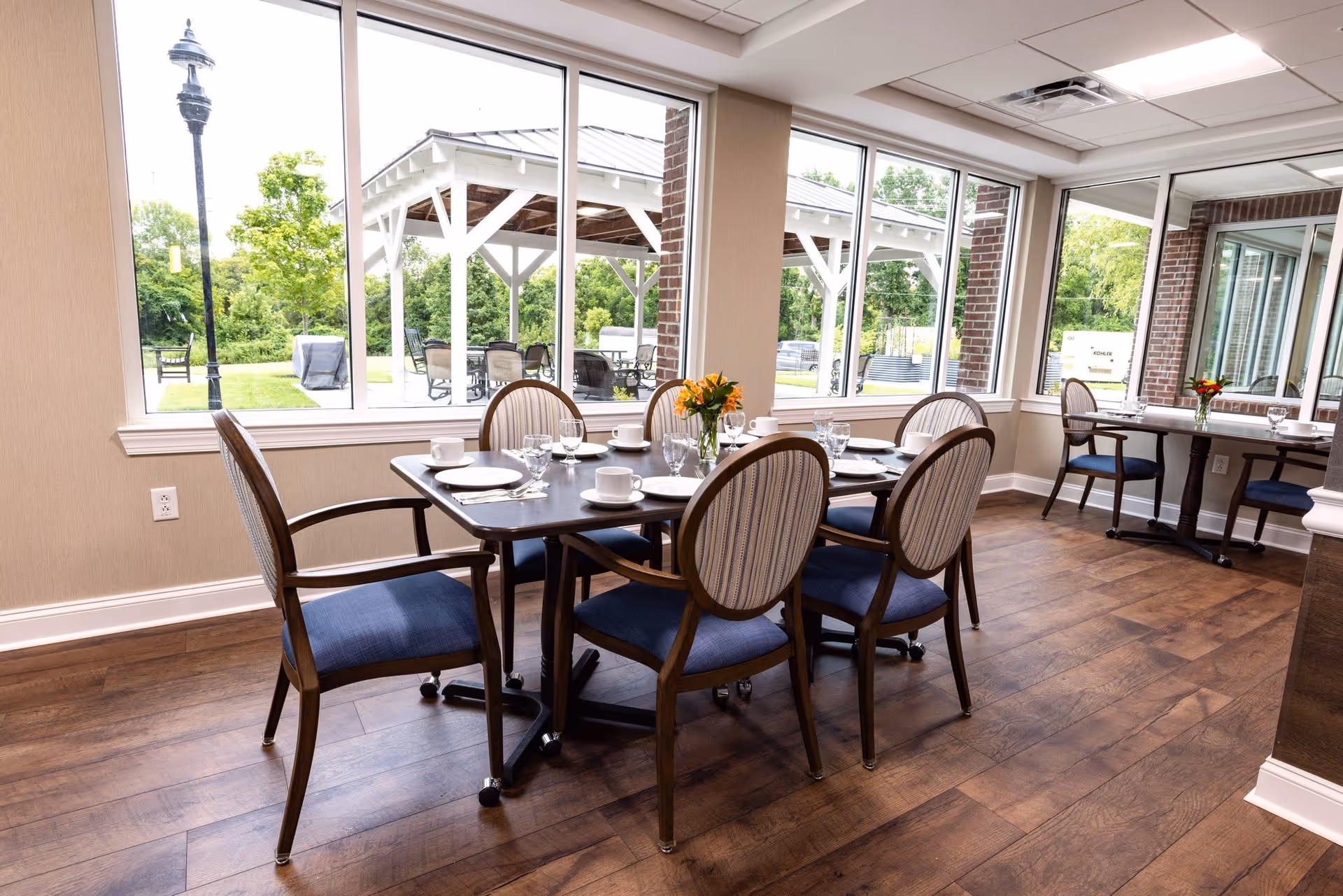 A bright dining room with wooden floors and large windows showing an outdoor patio area. The room has two dining tables set with plates, cups, glasses, and small flower arrangements. The chairs have wooden frames with blue cushioned seats and striped backs.