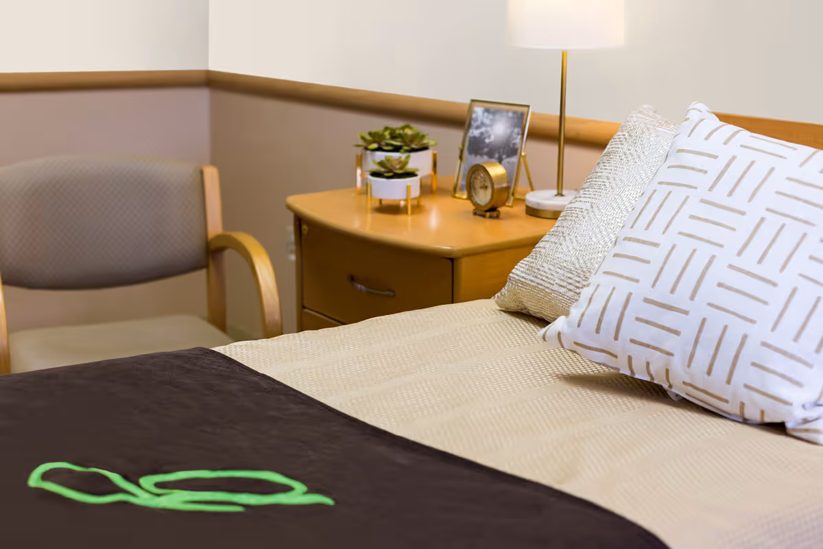 A neatly made bed with patterned pillows and a beige quilt in a room. Next to the bed is a wooden nightstand with a small lamp, a clock, a framed photo, and two small potted plants. A cushioned chair with wooden arms is positioned against the wall in the background.