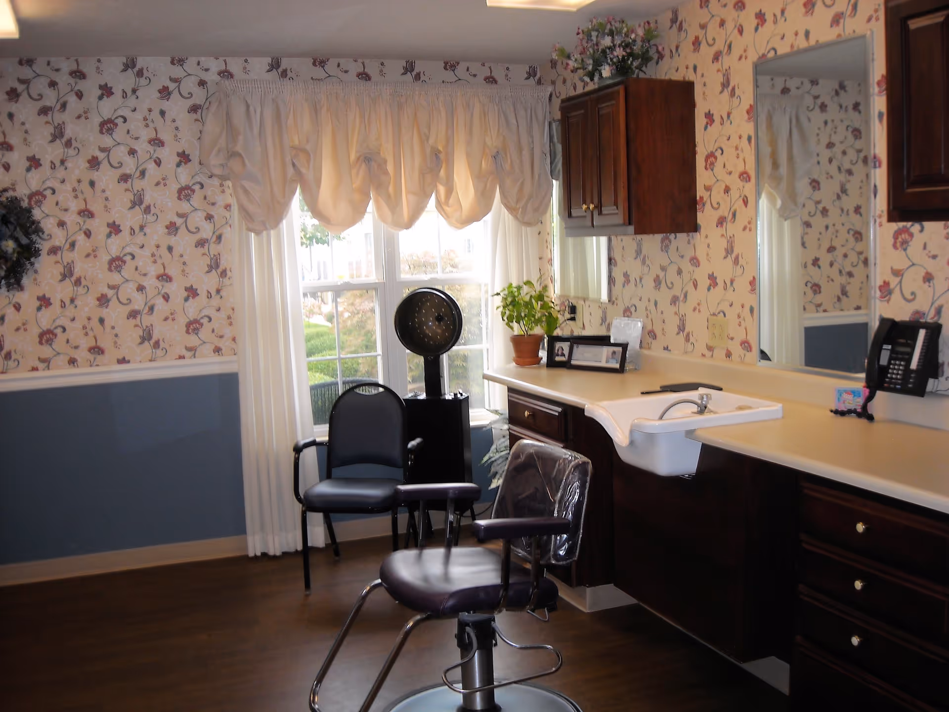 Interior of a salon or grooming area with floral wallpaper and a large window with white curtains. There is a salon chair with a plastic cover, a black chair, a hair dryer, a countertop with a sink, a potted plant, framed photos, and a wall-mounted telephone.