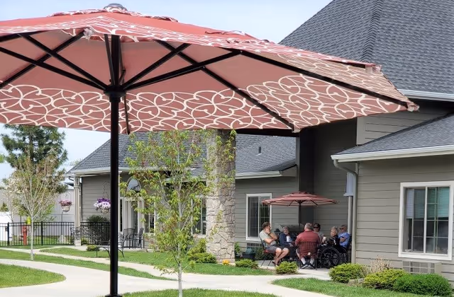 Outdoor patio area at The Renaissance At Coeur D'Alene with a large patterned umbrella in the foreground, a group of people sitting and conversing under a smaller umbrella near the building, surrounded by green grass, small trees, and shrubs.