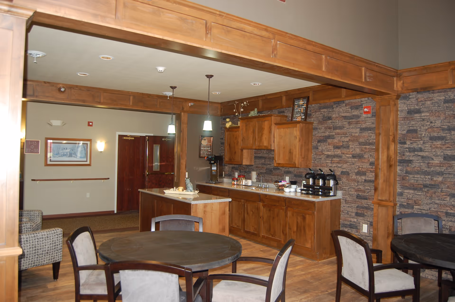 Interior view of a common area in StoneyBrook Suites Assisted Living featuring a small kitchen with wooden cabinets, a countertop with coffee dispensers, and a round table with chairs in the foreground. The walls have a stone-like texture and there are pendant lights hanging from the ceiling.