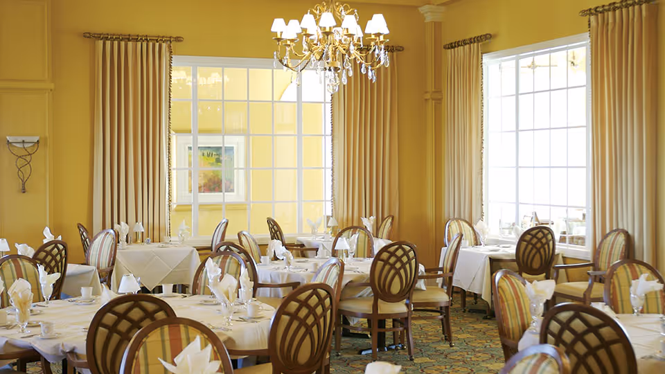 A bright and elegant dining room with multiple round tables covered in white tablecloths, each set with folded napkins, cups, and glasses. The room features large windows with beige curtains, yellow walls, a decorative chandelier hanging from the ceiling, and patterned chairs with wooden frames.