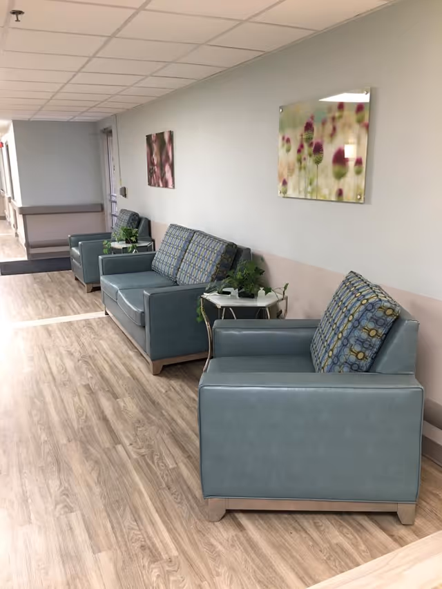 A hallway seating area with three blue cushioned chairs and two small tables with green plants. The walls are light-colored with two floral paintings hanging above the chairs. The floor has a wood-like finish and the ceiling has white tiles.