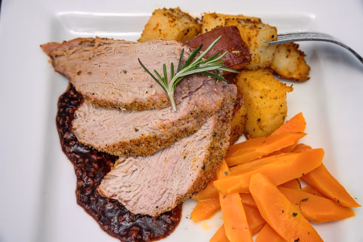 A plated meal featuring three slices of seasoned roast beef garnished with a sprig of rosemary, served with roasted potatoes and cooked carrot sticks on a white square plate.
