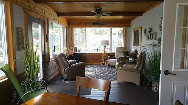Sunlit common living room with several recliner chairs, potted plants, a wooden ceiling, and large windows overlooking the outdoors.