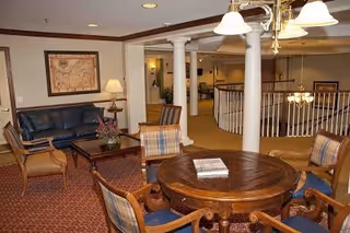 Interior view of a senior living facility lounge area with a round wooden table surrounded by four cushioned chairs. In the background, there is a seating area with a dark leather sofa, two wooden chairs, a coffee table with a flower arrangement, and a framed map on the wall. The space features carpeted flooring, white columns, and overhead lighting fixtures.