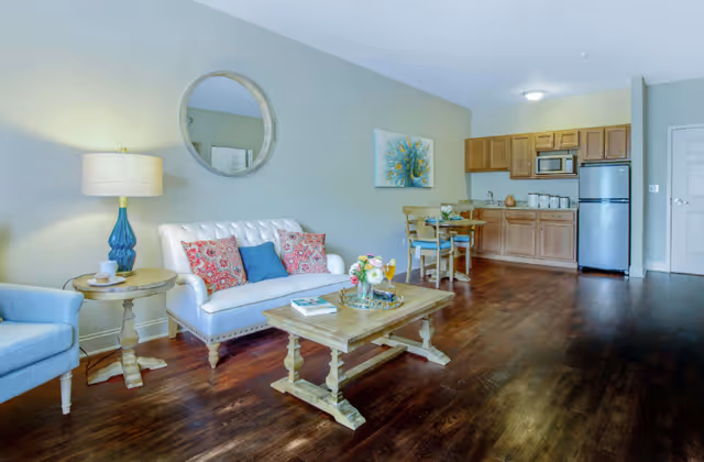 A bright and cozy living area featuring a white tufted sofa with colorful pillows, a wooden coffee table with a flower arrangement and books, a side table with a blue lamp, a round mirror on the wall, and a small dining table with two chairs. In the background, there is a kitchenette with wooden cabinets, a microwave, and a stainless steel refrigerator. The room has wooden flooring and light-colored walls.