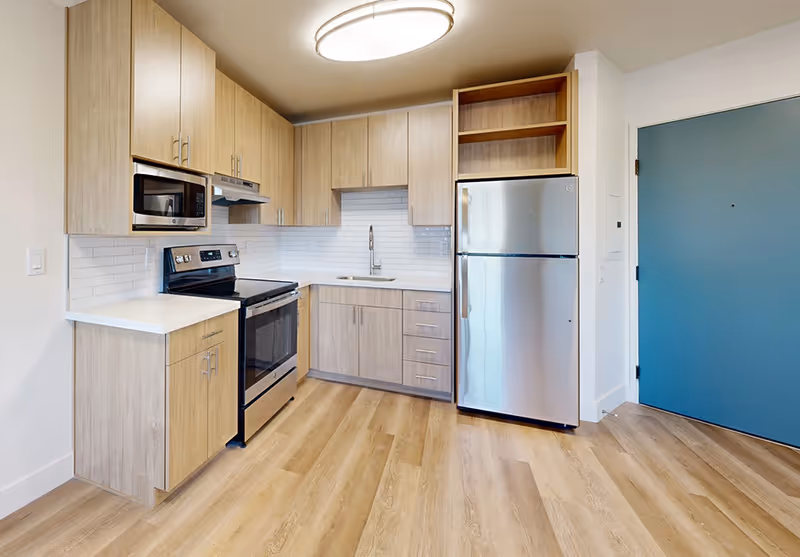 Modern kitchen with light wood cabinets, stainless steel refrigerator, stove, and microwave. White countertops and white subway tile backsplash. Light wood flooring and a blue door to the right.