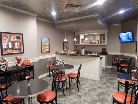 A cozy dining area with several round tables and red cushioned chairs. The room features a small kitchen counter with coffee and snacks, pendant lights hanging from the ceiling, and a wall-mounted TV. The walls are decorated with framed pictures and a menu board. The floor has a gray tile pattern, and the ceiling has a decorative metallic finish.