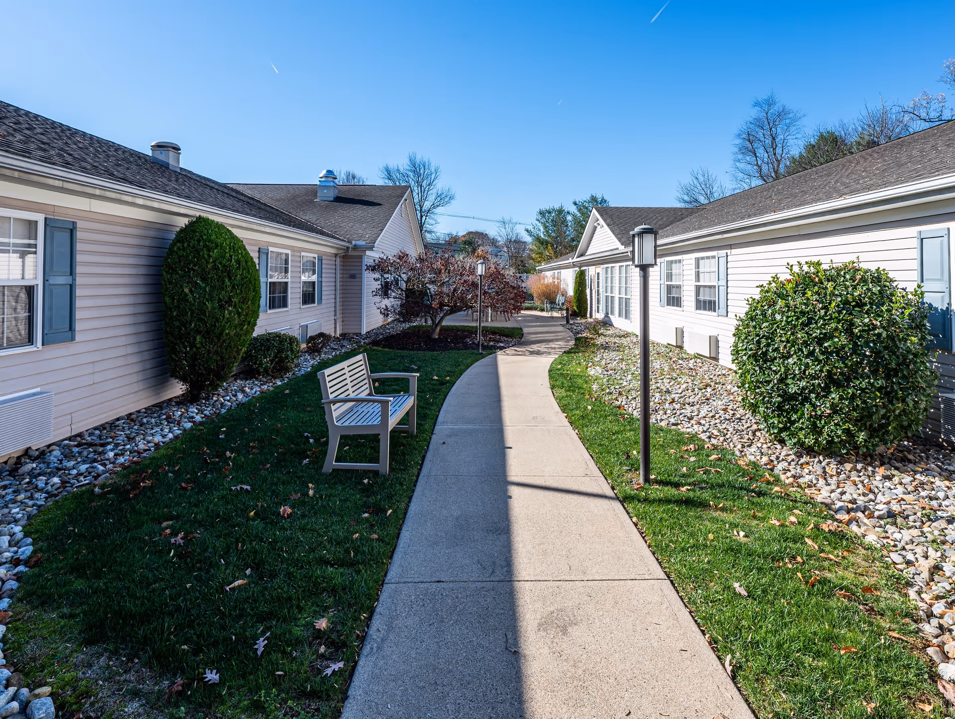 Outdoor pathway between two single-story buildings with beige siding and blue shutters. The pathway is flanked by green grass, shrubs, and small trees. There is a wooden bench on the left side and lamp posts along the path under a clear blue sky.