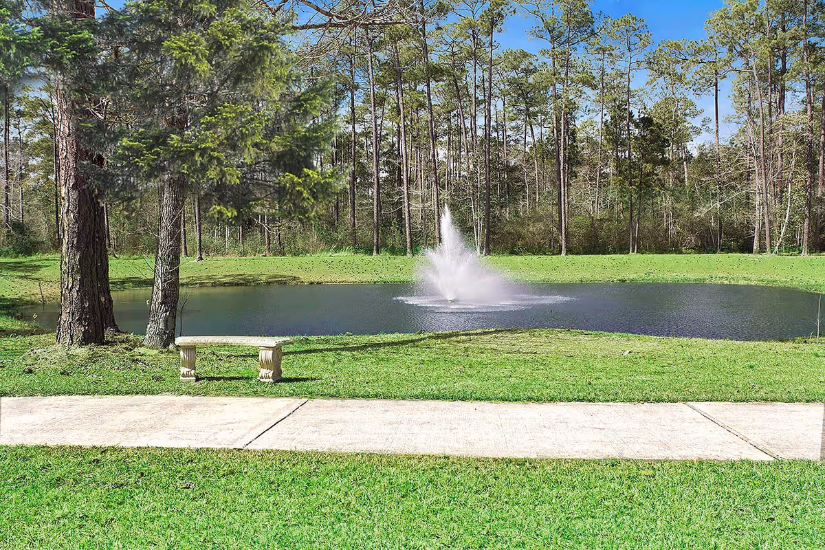A peaceful outdoor scene at Summerfield Senior Living featuring a small pond with a water fountain in the center, surrounded by green grass and tall pine trees. A stone bench is placed near the pond along a concrete pathway under a clear blue sky.