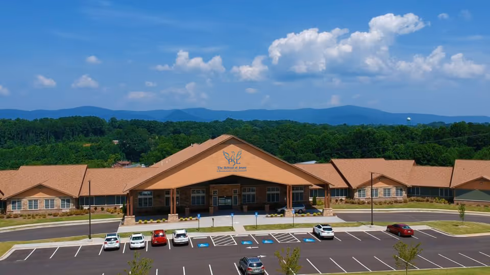 Front view of The Retreat at Jasper senior living building with a parking lot, surrounding trees, and mountains under a blue sky.