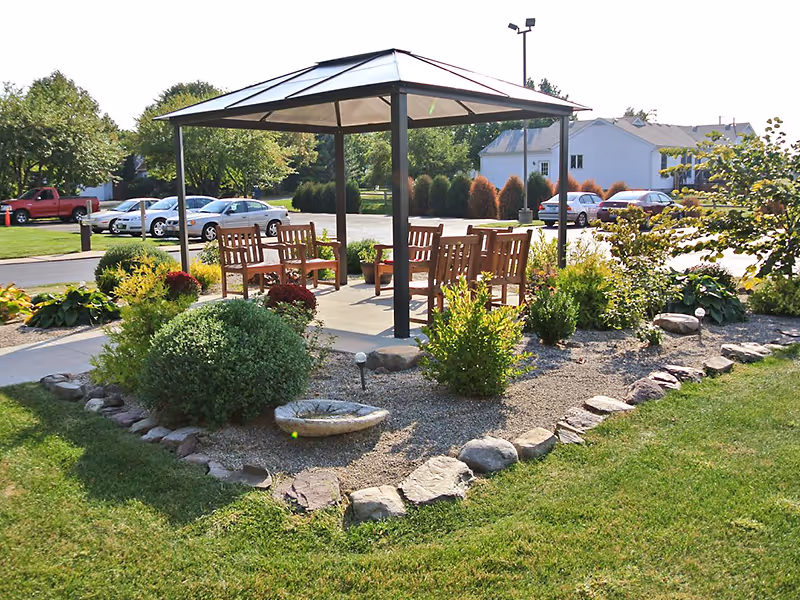 Outdoor seating area with a metal gazebo covering wooden chairs and a table, surrounded by landscaped garden with bushes, rocks, and a birdbath, with parked cars and houses visible in the background.